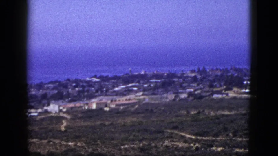 1953 California 8mm frame showing a coastal settlement below an elevated viewpoint, with open hillsides and the Pacific Ocean in the distance.