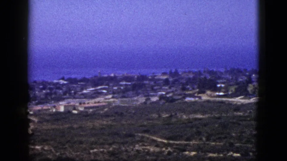 1953 California coastal view with scattered buildings, hillside terrain, pale roads, and the Pacific Ocean beyond the town.