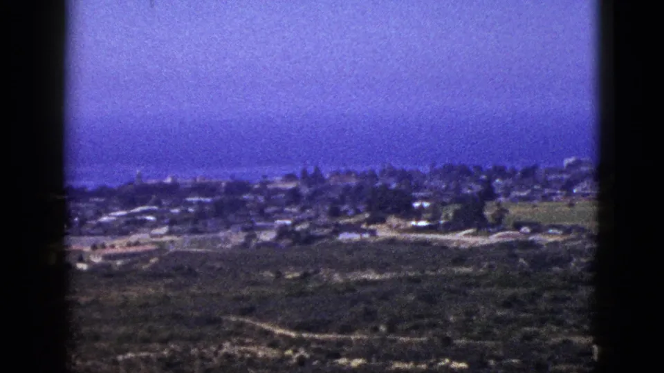 1953 California home-movie frame showing a coastal town across open land, with soft grain, dark border, and ocean behind the settlement.