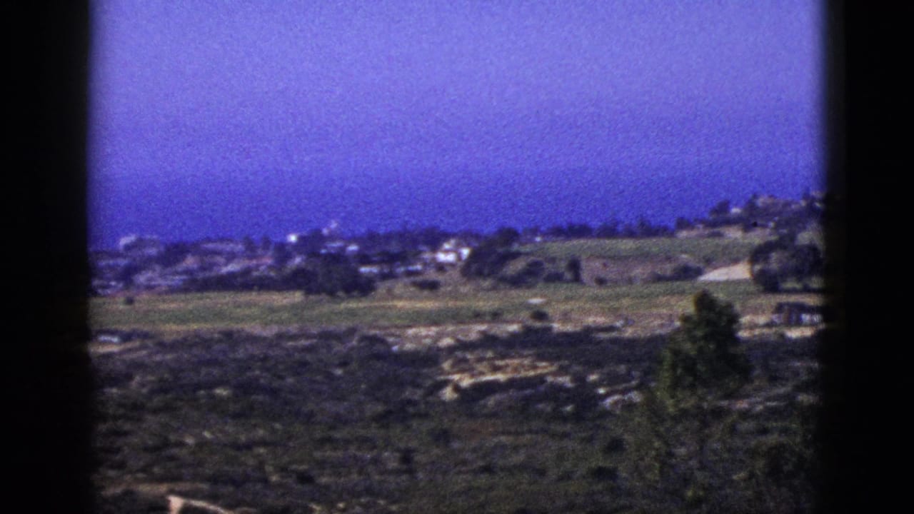 1953 California 8mm frame showing open hills, distant houses, trees, rocky ground, and a deep blue-purple sky.