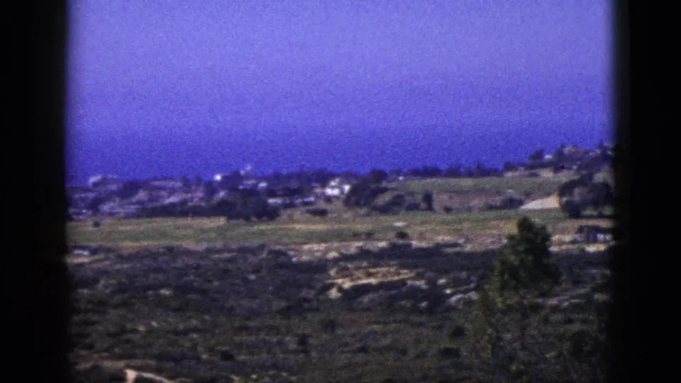 1953 California 8mm frame showing open hills, distant houses, trees, rocky ground, and a deep blue-purple sky.