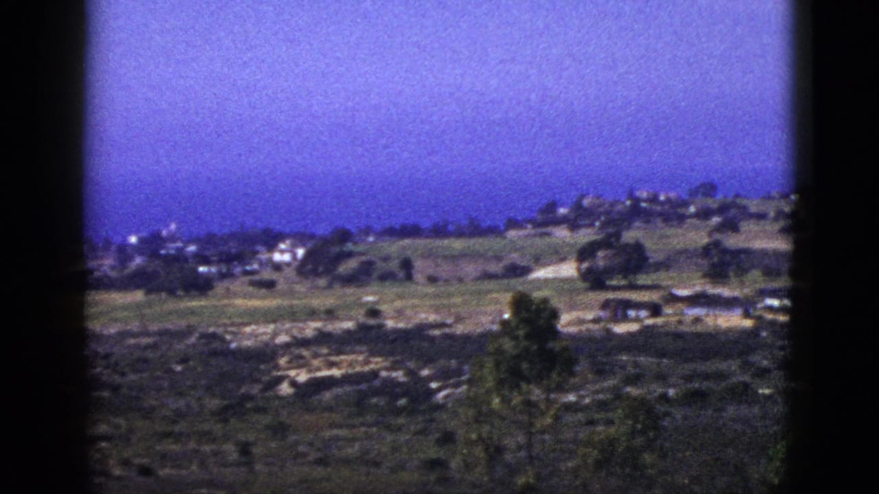 Open hillside landscape with distant homes, trees, foreground scrub, and dark film borders.