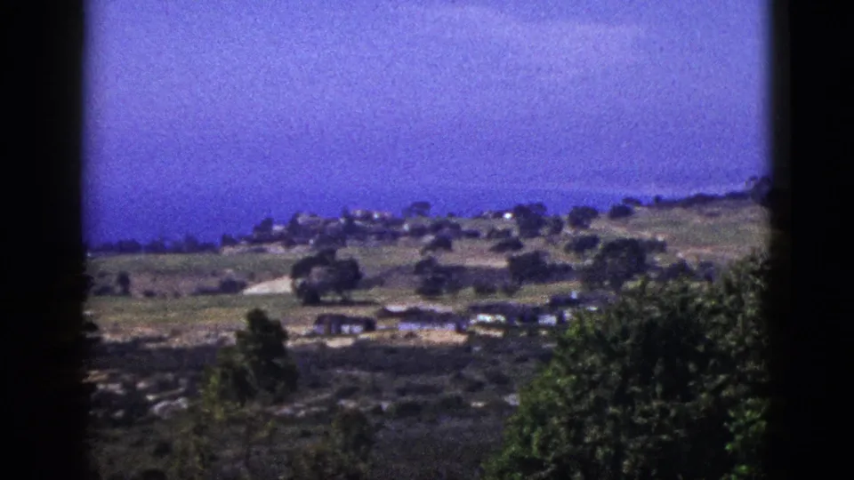 1953 California 8mm view of a hillside with pale road, small buildings, trees, foreground foliage, and possible coastline beyond.