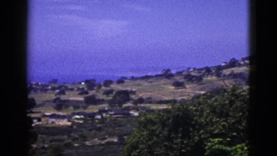 1953 California home-movie frame showing distant hills, clustered houses, foreground foliage, and a blue-purple sky.