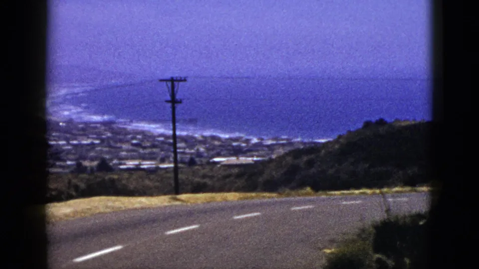 1953 California 8mm frame showing a paved coastal road, roadside grass, utility pole, distant homes, and the blue ocean beyond.