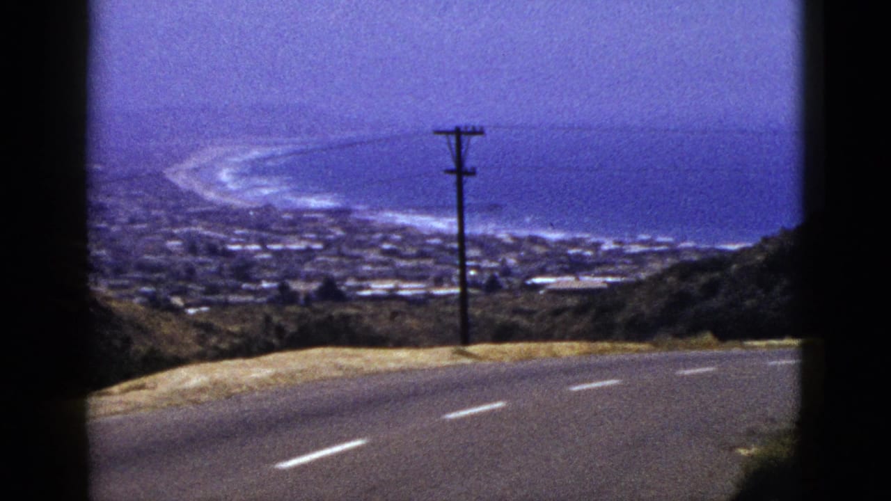 1961 California 8mm frame showing a curving hillside road, utility pole, distant coastal town, surf line, and open ocean.