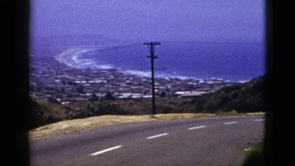 1961 California 8mm frame showing a curving hillside road, utility pole, distant coastal town, surf line, and open ocean.