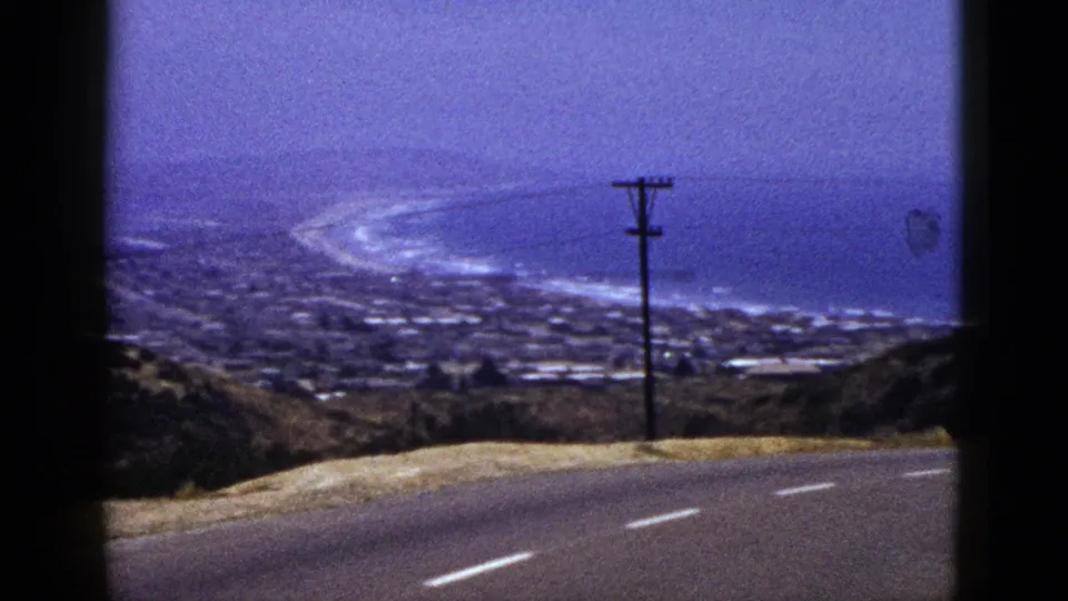 1961 California 8mm frame with a utility pole, sloping roadside, distant shoreline, beach surf, and blue ocean under haze.