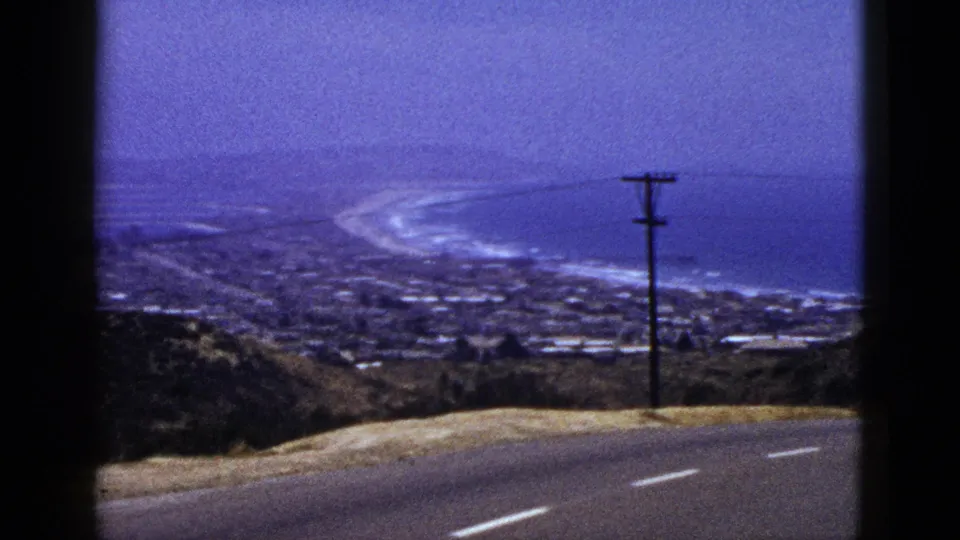 1961 California 8mm frame looking past a roadside slope and utility pole toward a coastal town, shoreline, and open water.