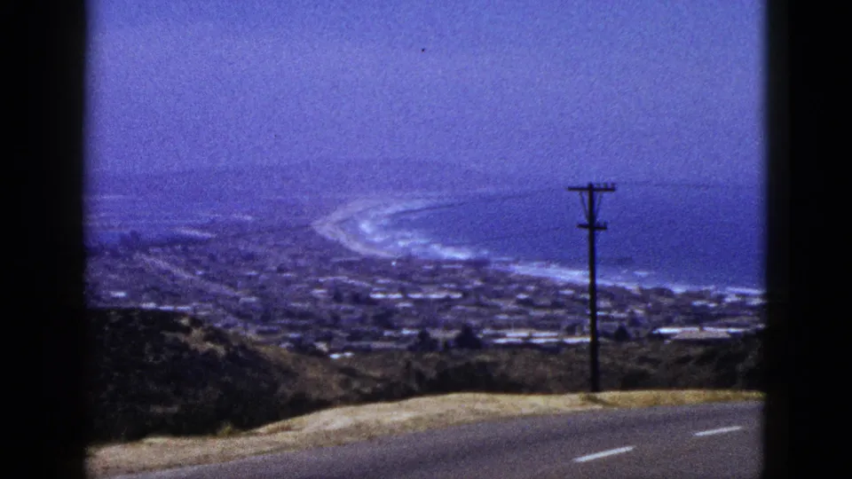 1961 California 8mm frame showing a coastal panorama from a road, with a utility pole, distant buildings, surf, and hazy ocean.