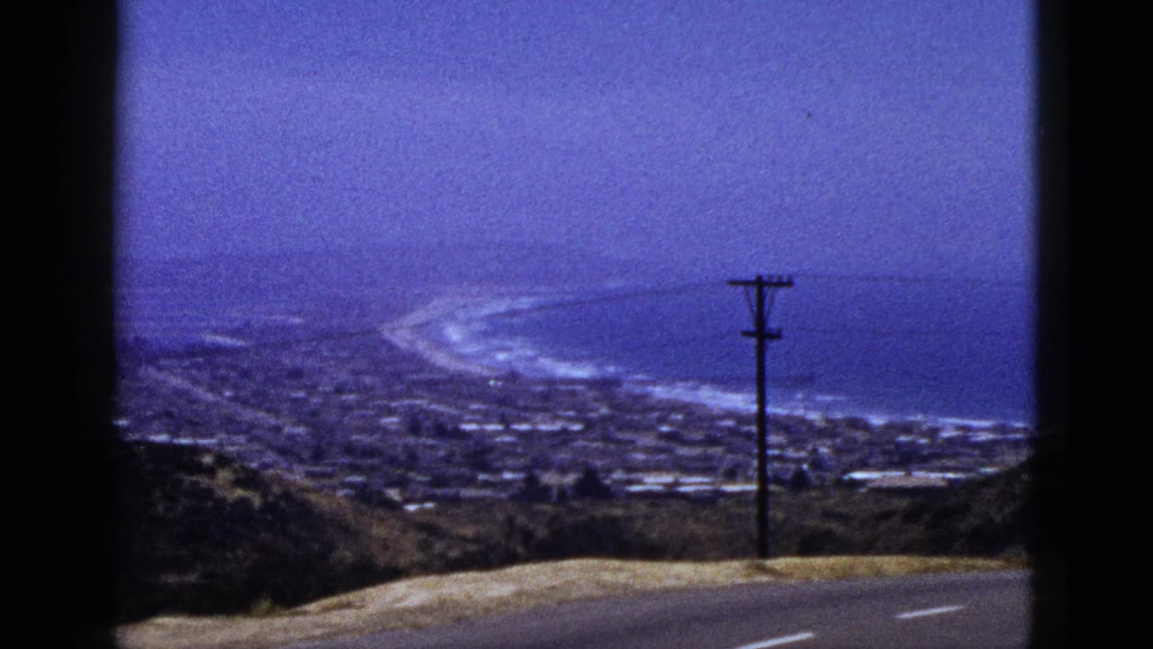 1953 California home-movie frame showing a coastal town below, surf along a curved beach, hillside foreground, and a roadside utility pole.