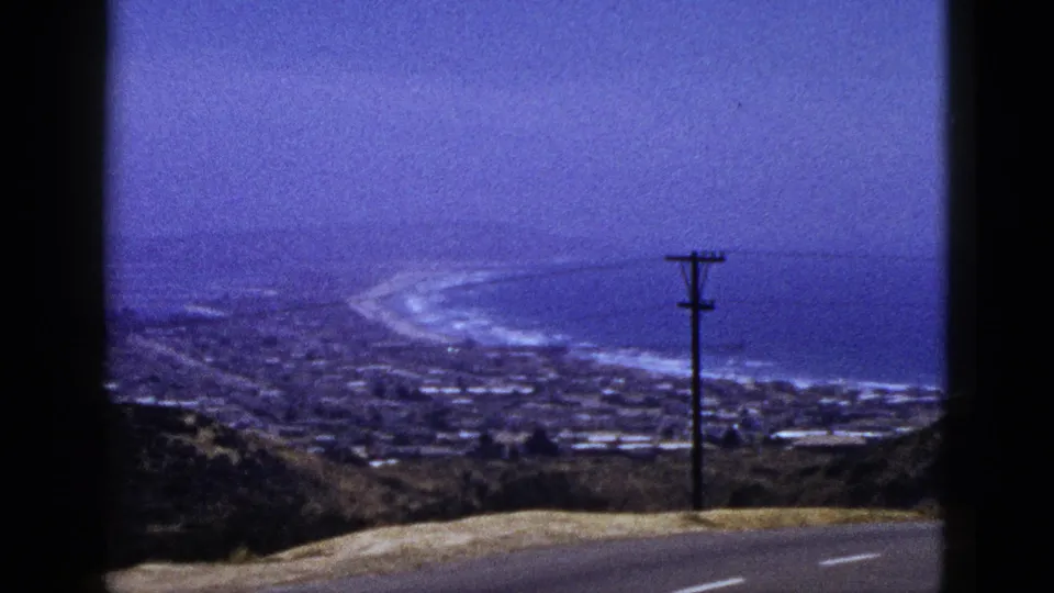 1953 California home-movie frame showing a coastal town below, surf along a curved beach, hillside foreground, and a roadside utility pole.