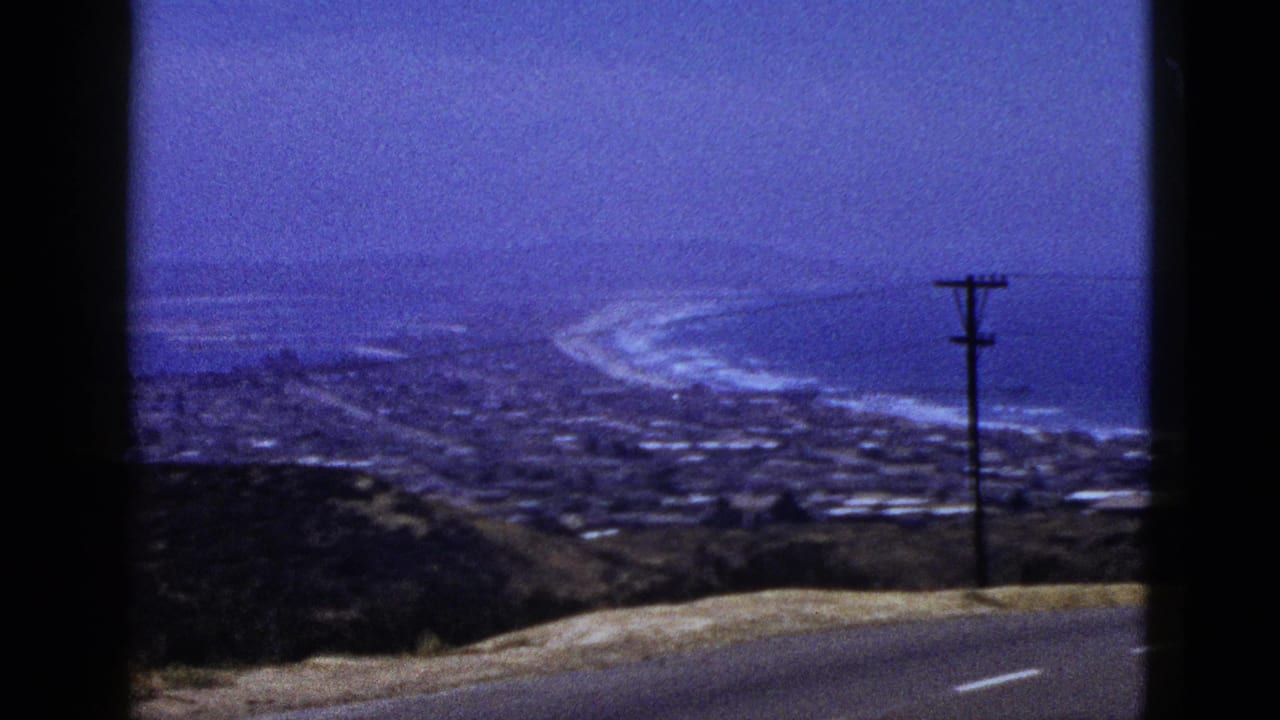 Roadside panorama of a coastal settlement, beach curve, ocean water, and a utility pole near the right edge.