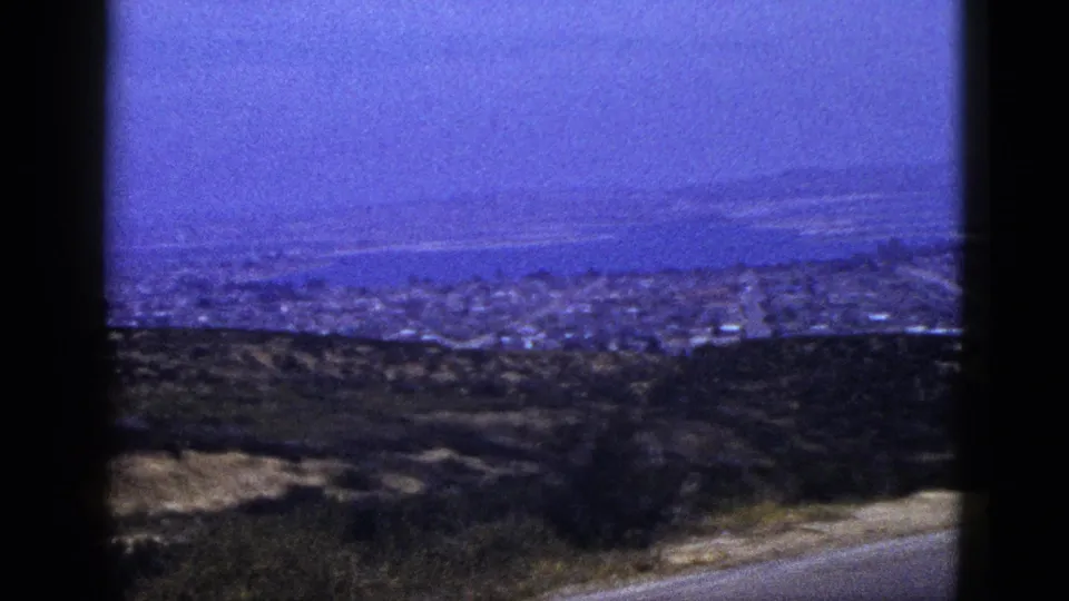Soft 8mm frame from 1953 showing a hazy California coastal town below a blue bay, seen from a hillside roadside viewpoint.