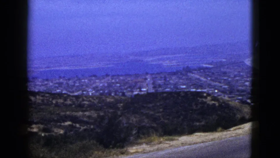 1953 California home-movie frame looking from a roadside hillside across brush and town rooftops toward a broad blue bay.