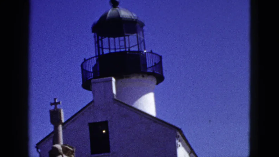 California 1953 home-movie frame showing the lighthouse-like tower, adjacent white building, cross, and dark window under a clear sky.