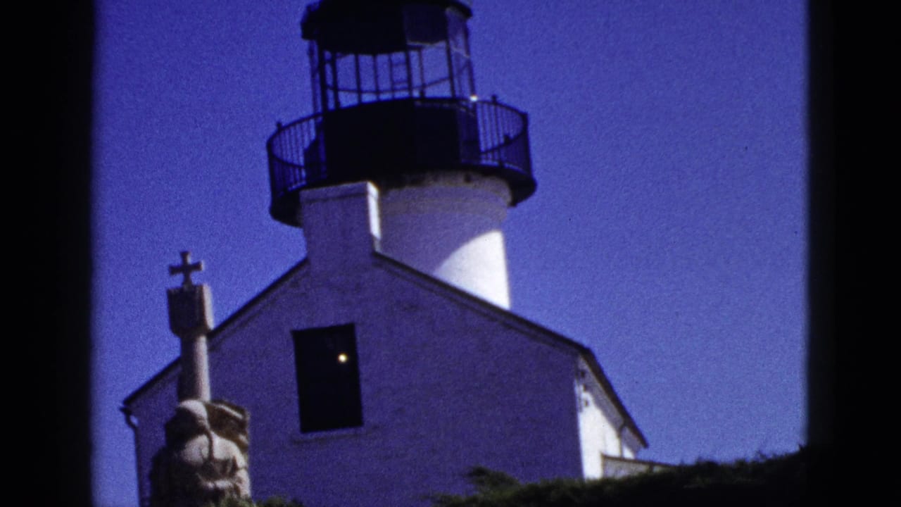 1953 California 8mm frame of a white lighthouse-like building with a black lantern room, dark window, stone robed statue, foliage, and blue sky.