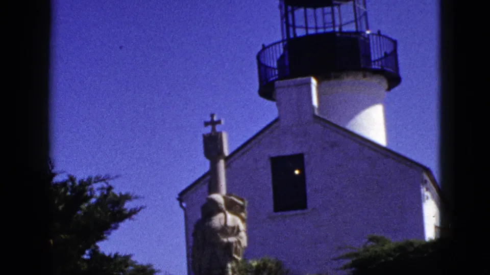 1953 California 8mm frame closer on the white lighthouse structure, black lantern railing, dark window, robed statue with cross, foliage, and saturated sky.