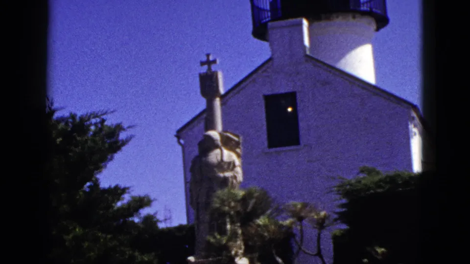 1953 California 8mm frame showing the white lighthouse and attached building, black railing, robed statue with cross, shrubs, dark window, and blue sky.