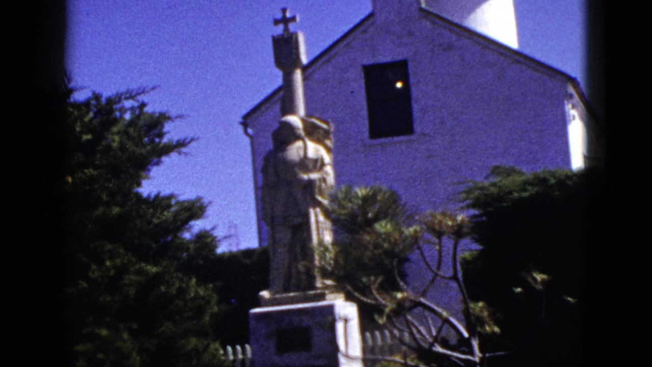 1953 California 8mm frame showing a robed stone statue with a cross near a white church wall, trees, and blue sky.