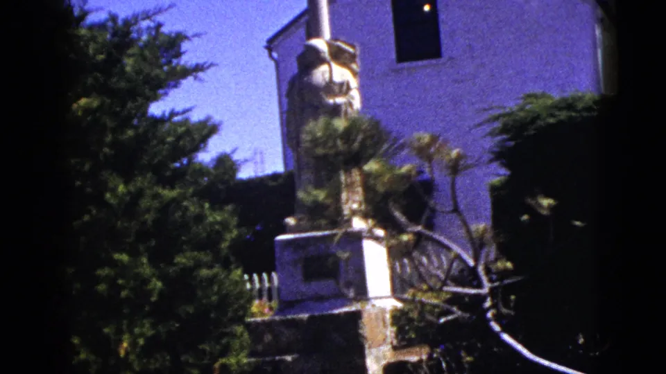 1953 California home-movie view of the religious statue and pedestal framed by greenery beside a white church facade.