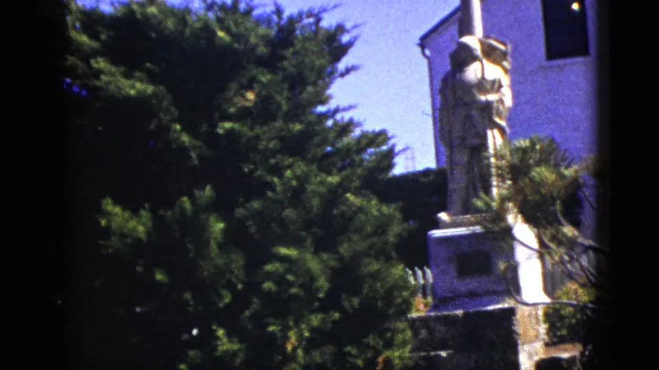 1953 California 8mm frame with the robed statue partly obscured by shrubs near the white church structure.