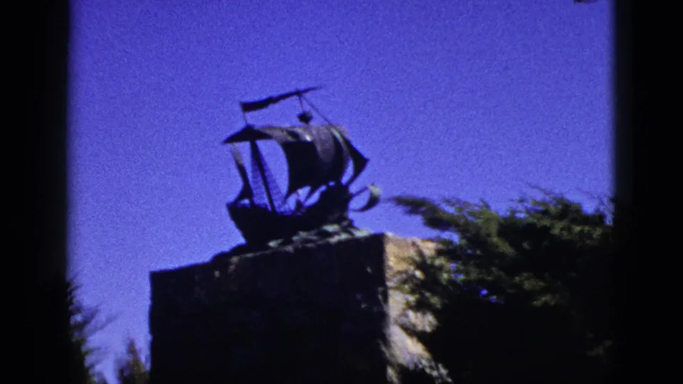 1953 California 8mm frame showing a nautical ship sculpture on a pedestal against clear sky, with shrubs at the edge.