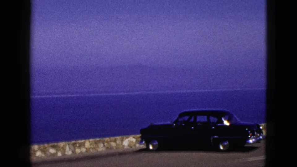 1953 California coastal road frame with a dark mid-century sedan near a low stone wall and ocean horizon.