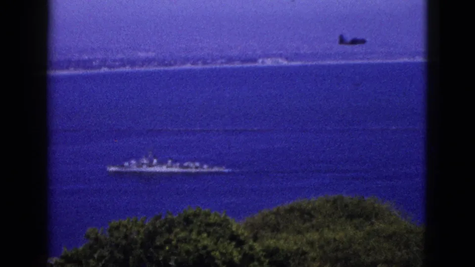1953 California coastal frame with a distant aircraft above water and a naval-looking ship offshore.