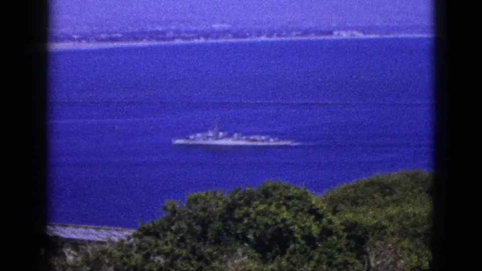 1953 California 8mm frame showing a naval-looking ship offshore beyond coastal vegetation.
