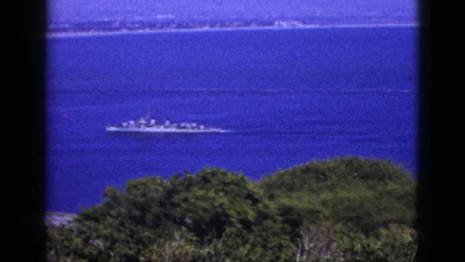 1953 California coastal view with a distant naval-style ship on blue water, framed by trees and film gate edges.