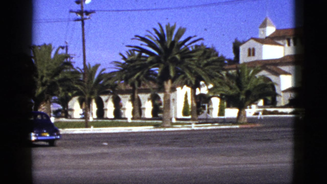 White Spanish-style building behind palm trees, with a dark car at the street edge.