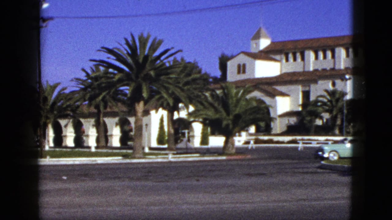 White building with palms and a pale car near the right side of the street.