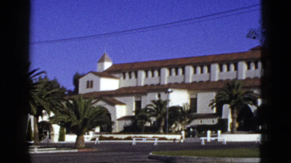 1953 California view of a long white building with red tile roof, arched entry, palms, white fence, and empty roadway.
