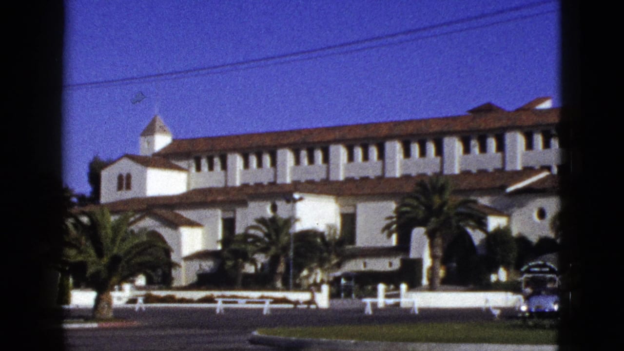 White stucco Spanish Colonial Revival building with red tile roof, palms, and cars along the street.