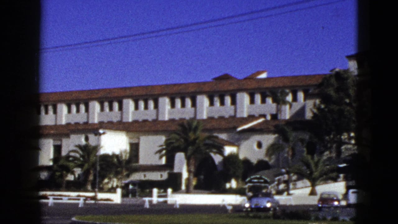 Long white stucco building with red roof, palms, circular window, and cars near the curb.