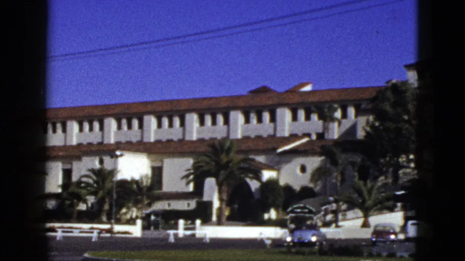 1953 California exterior view of a Spanish Colonial Revival style building with palms, a circular window, and cars on the street.