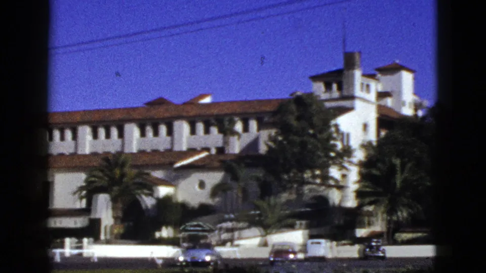 1953 California 8mm frame with a partial Spanish Colonial Revival facade, tower section, palms, and period cars along the street.
