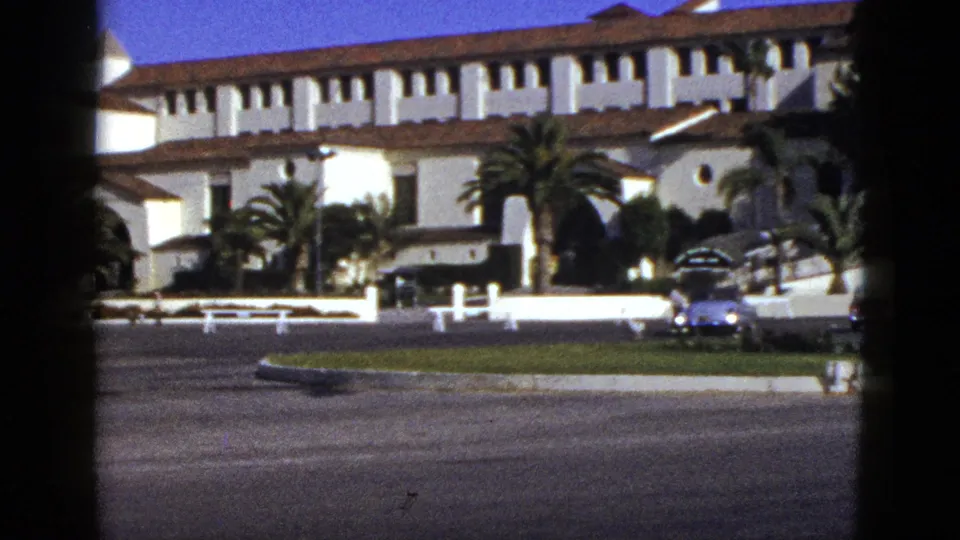 1953 California street-level view of a Spanish Colonial Revival building, palms, broad pavement, and a blue early-1950s car.
