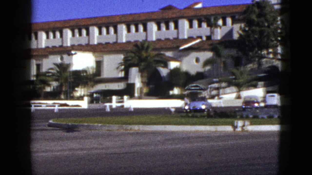 1953 California home-movie frame showing a Spanish Colonial Revival-style exterior, palm trees, a low wall, and mid-century cars across a road.