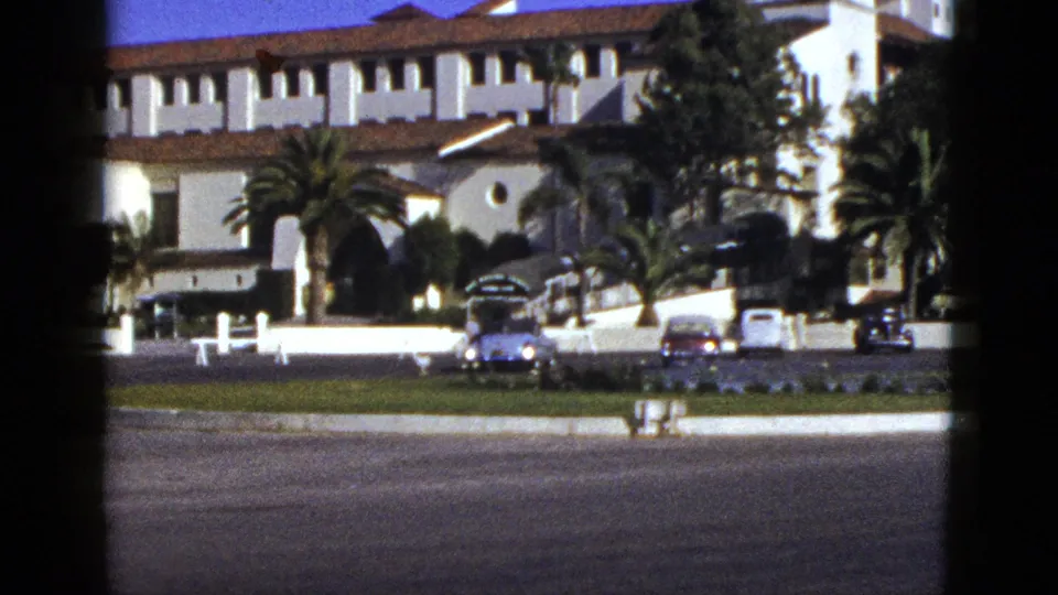 1953 California street view of a large stucco building with red tile roofing, palms, a low wall, lawn, and parked mid-century vehicles.
