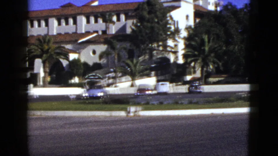 1953 California home-movie frame with a closer view of the stucco building, red roofline, palms, parked cars, and roadway foreground.