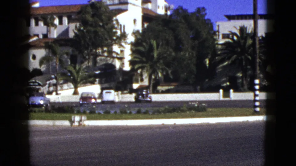 1953 California frame showing the building partly obscured by trees, with palms, parked cars, a white wall, and broad road in front.