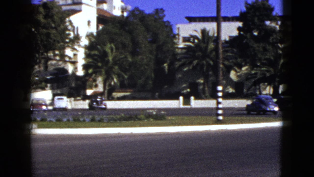 1953 California 8mm frame showing cars on a broad road, palms, lawn, and a white Spanish-style building beyond.