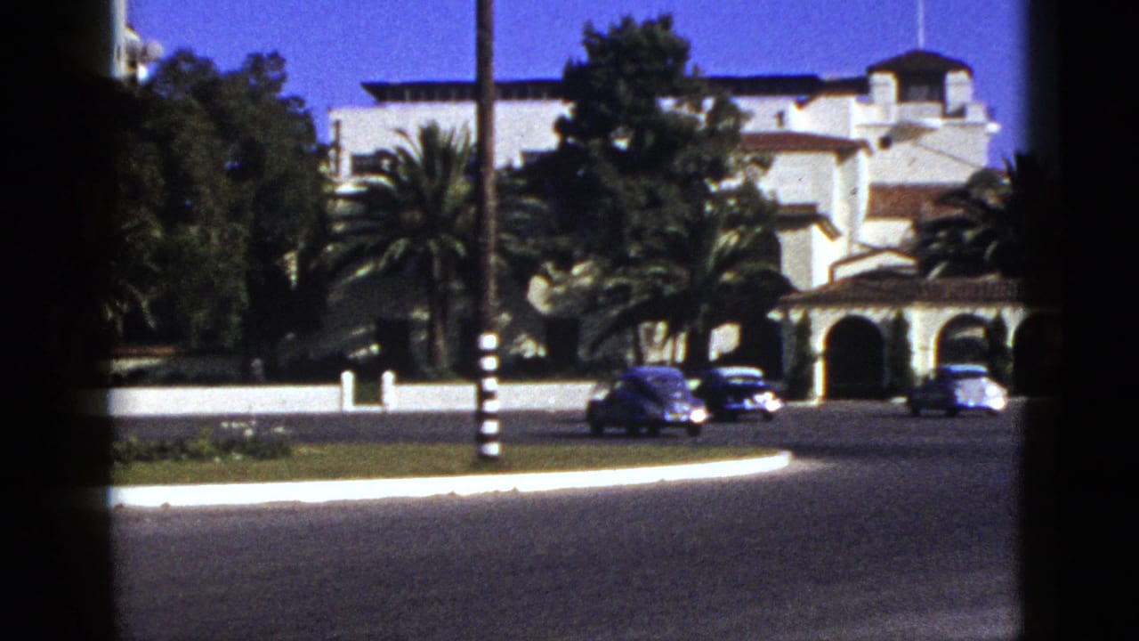 Cars drive past palms and arched building entrances on a sunny street.