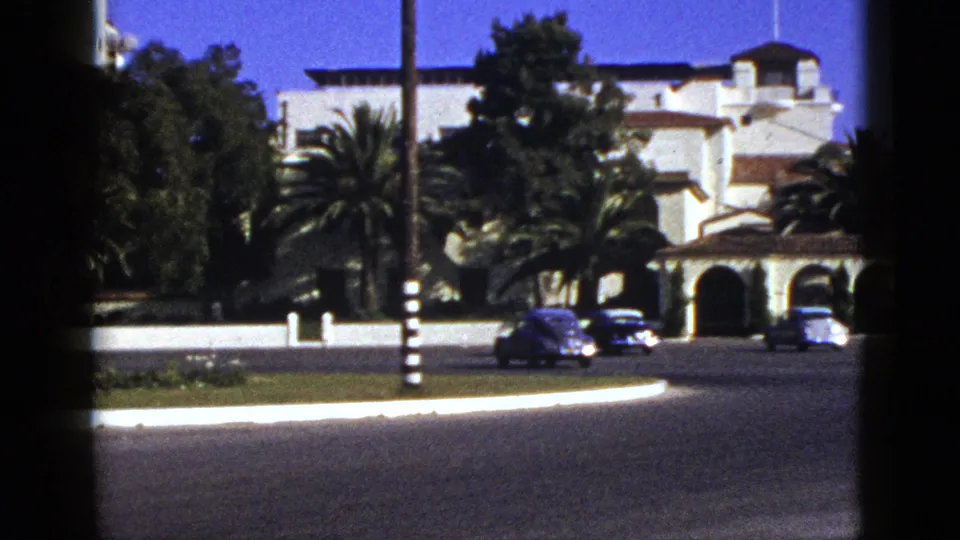 1953 California street view with early-1950s cars, palm trees, and a Spanish-style building with arched entryways.