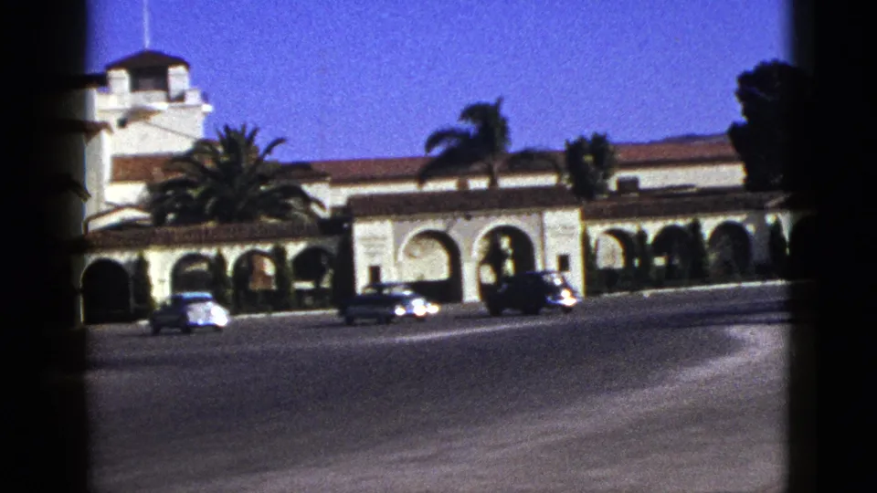 1953 California frame with an arched Spanish-style facade, red-tile roof, palms, and cars crossing the foreground road.