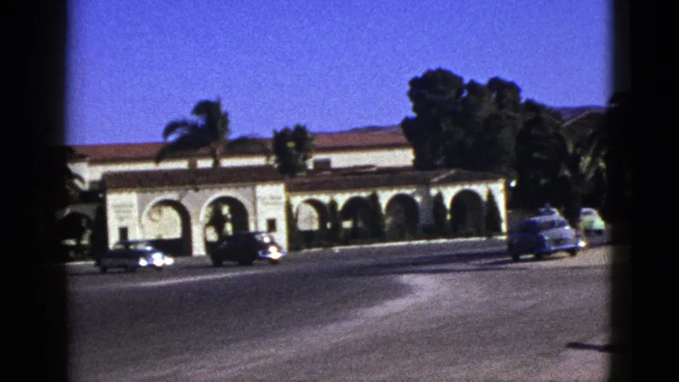 1953 California 8mm view of a broad street, classic cars, palms, and a Spanish-style building with repeating arches.