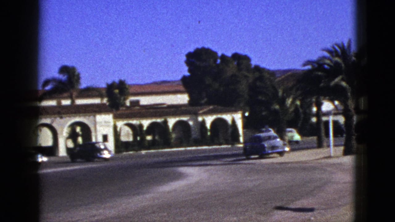 Vintage cars passing a Spanish-style arched building beside palm trees.
