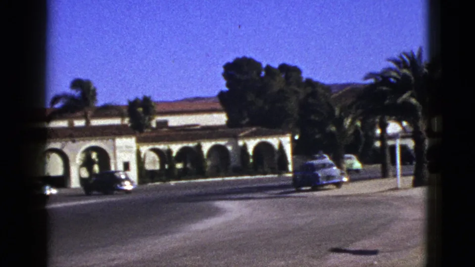 1953 California 8mm frame showing vintage cars on a road beside an arched Spanish-style building and palms.